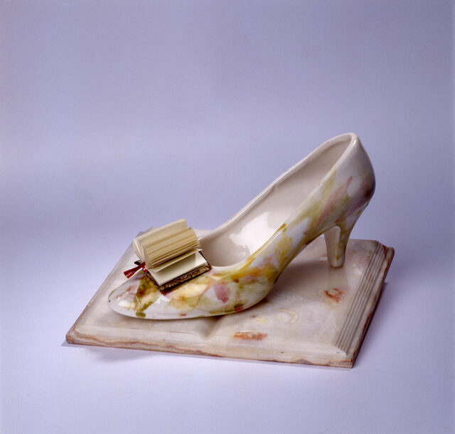 A woman's glass slipper rests on the open pages of a porcelain book. In place of a bow on the toe of the slipper is a paper book with pages fanned out.