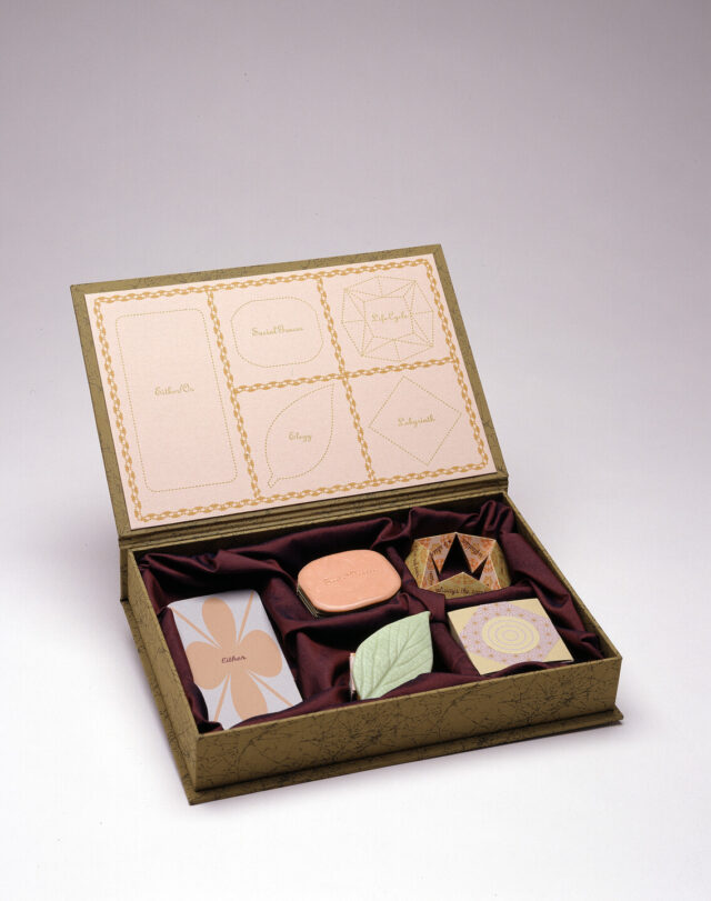 A green-gold box with its attached lid flipped open holds five objects shaped like candies resting on burgundy fabric. The inside of the lid displays outlines of each object, with the words 