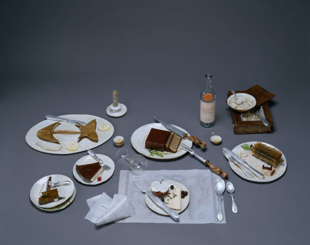 A photo of a still life of the remnants of a large meal. The half-eaten food is actually made of books that are sculpted to resemble fishbones, slices of pie, and other food scraps.