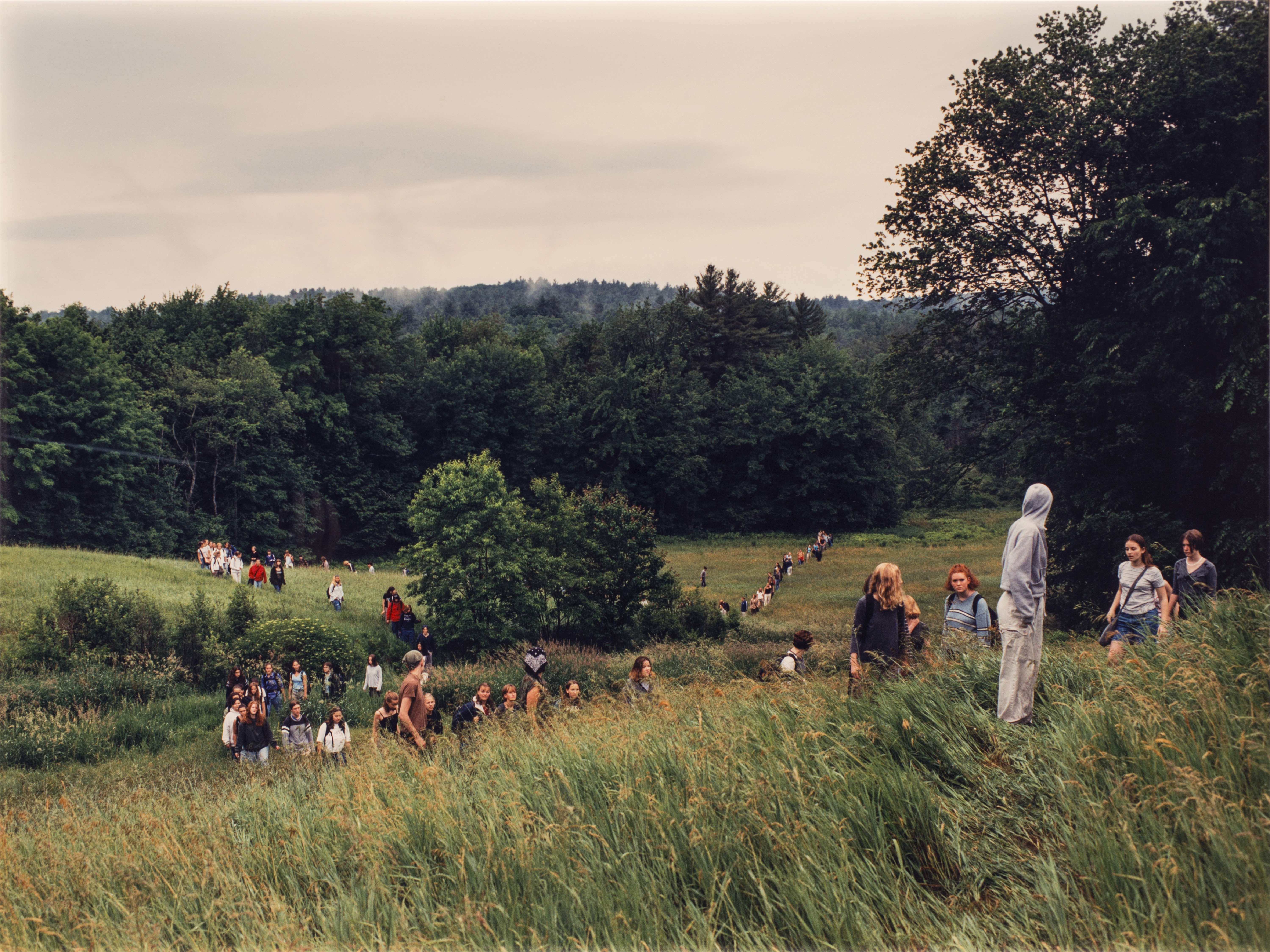 A landscape photograph featuring a field of tall green grass surrounded by leafy trees and a grey sky. A large group of people of varying ages traverse the field on foot. In the background the people form two lines, and a smaller group is congregated in the foreground.
