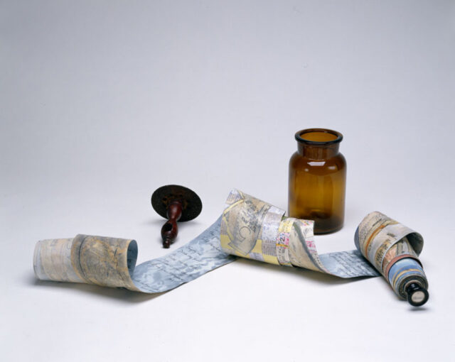 A small antique amber jar and a wooden scroll holder are arranged alongside a long scrolled paper with handwriting on one side and the faint hint of collaged sewing patterns, maps, and newsprint on the reverse side.