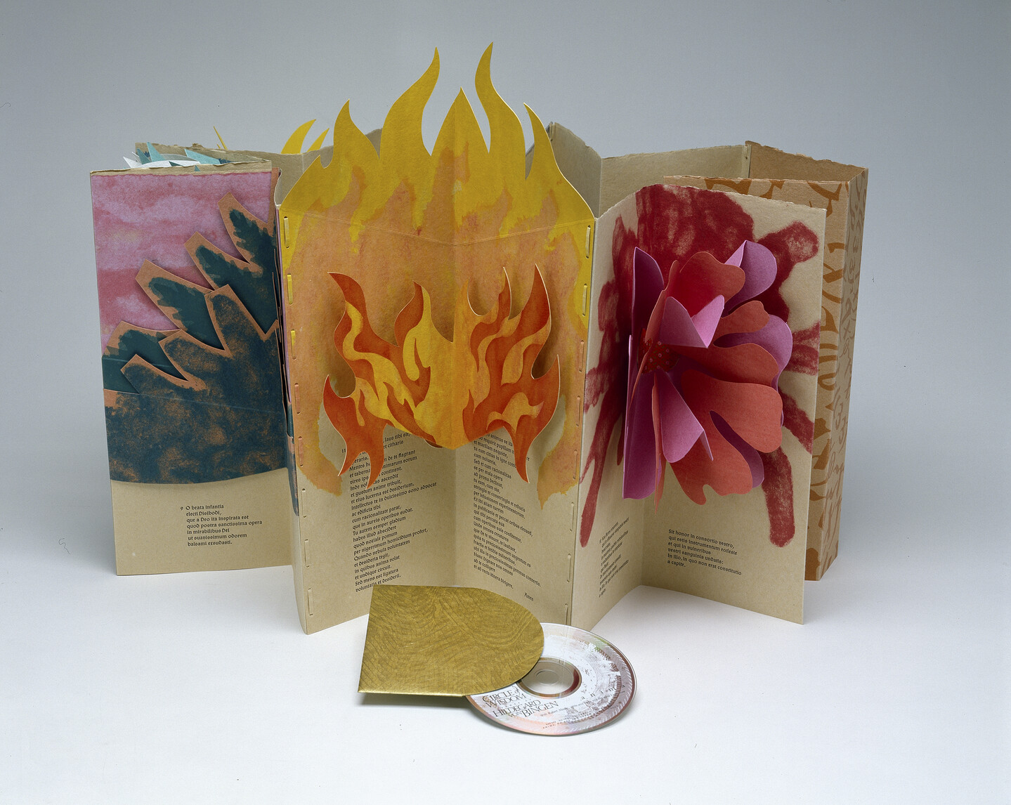 An accordion-style pop-up book made of paper displayed upright and open. The central portion has flames made of paper that extend beyond the top edge of the book. In front of the book there is a CD coming out of its golden holding case.