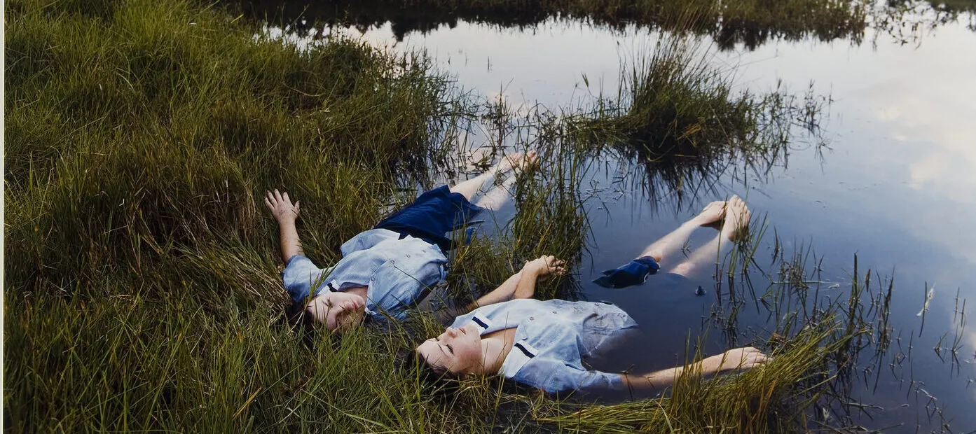 Two adolescent girls with light skin wearing matching blue uniforms float side-by-side on the edge of a grassy pond while holding hands.