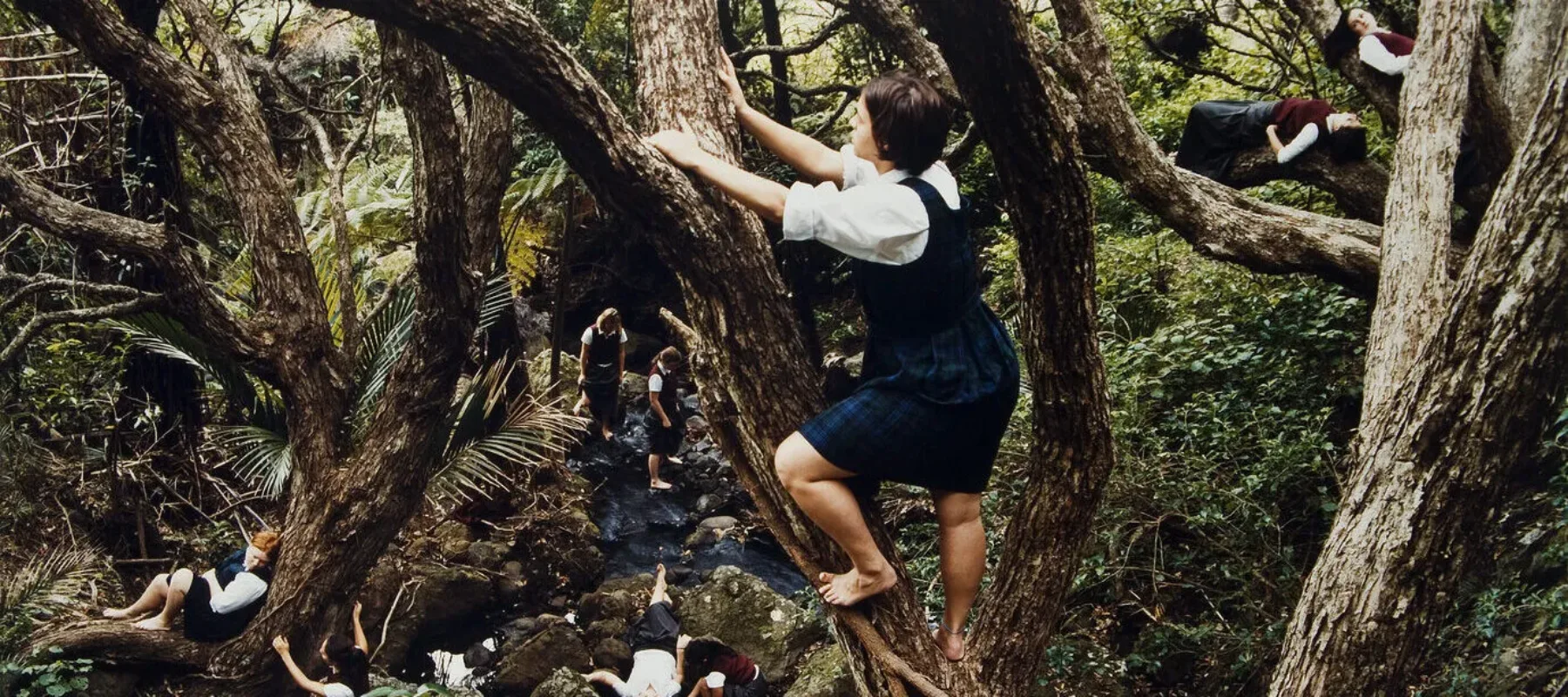 Color photograph of a dense forest with a light skin-toned girl with bare feet climbing a tree at center. Below her to the right is another light skinned girl sitting against the trunk of a tree. Other figures are scattered throughout the forest in the background.