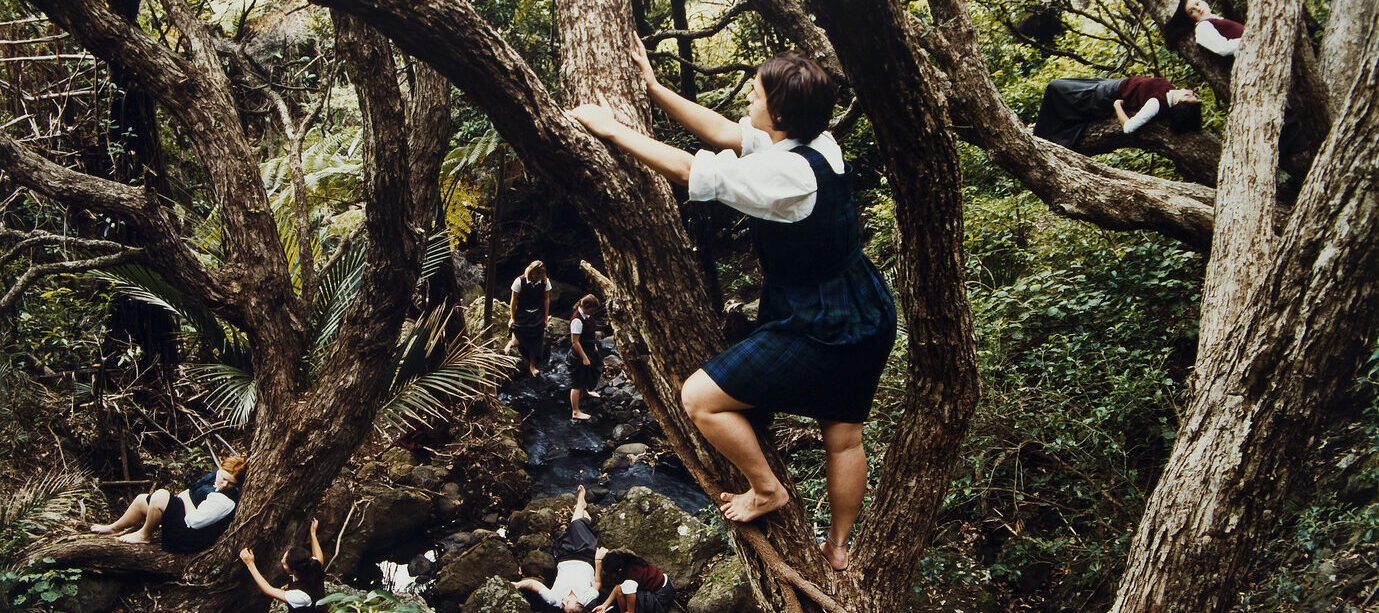 Color photograph of a dense forest with a light skin-toned girl with bare feet climbing a tree at center. Below her to the right is another light skinned girl sitting against the trunk of a tree. Other figures are scattered throughout the forest in the background.