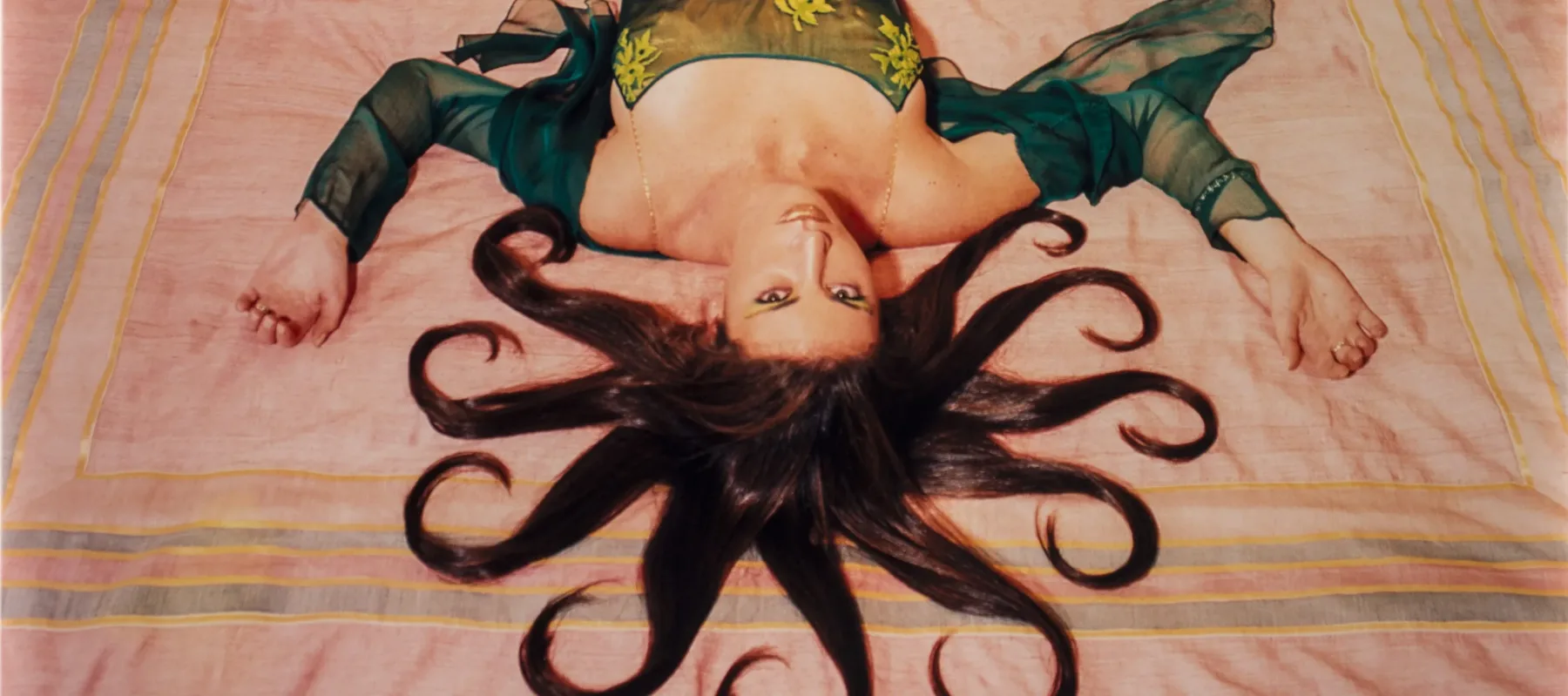 A slim, light-skinned woman wearing a long, emerald-green dress gazes up at the viewer from the foot of the bed, her bare feet near the headboard. Brown hair radiates from her head in 10 curled, snake-like segments. Her arms, bent at the elbows, extend from her body to form a W.