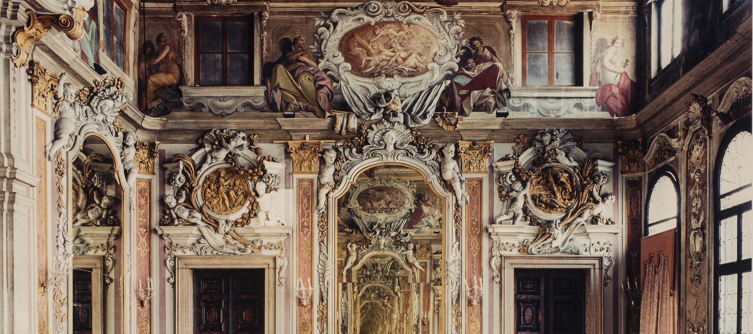 A color photograph of a large, ornate room in a palace. The expansive floor is bare and empty. At the other end of the room, a large ornate mirror hangs between two dark wooden doors. The moldings and details above the doors and mirror are extravagantly designed and a dramatic mural is featured at the very top of the wall. In the mirror, a small figure is seen, seemingly the photographer.