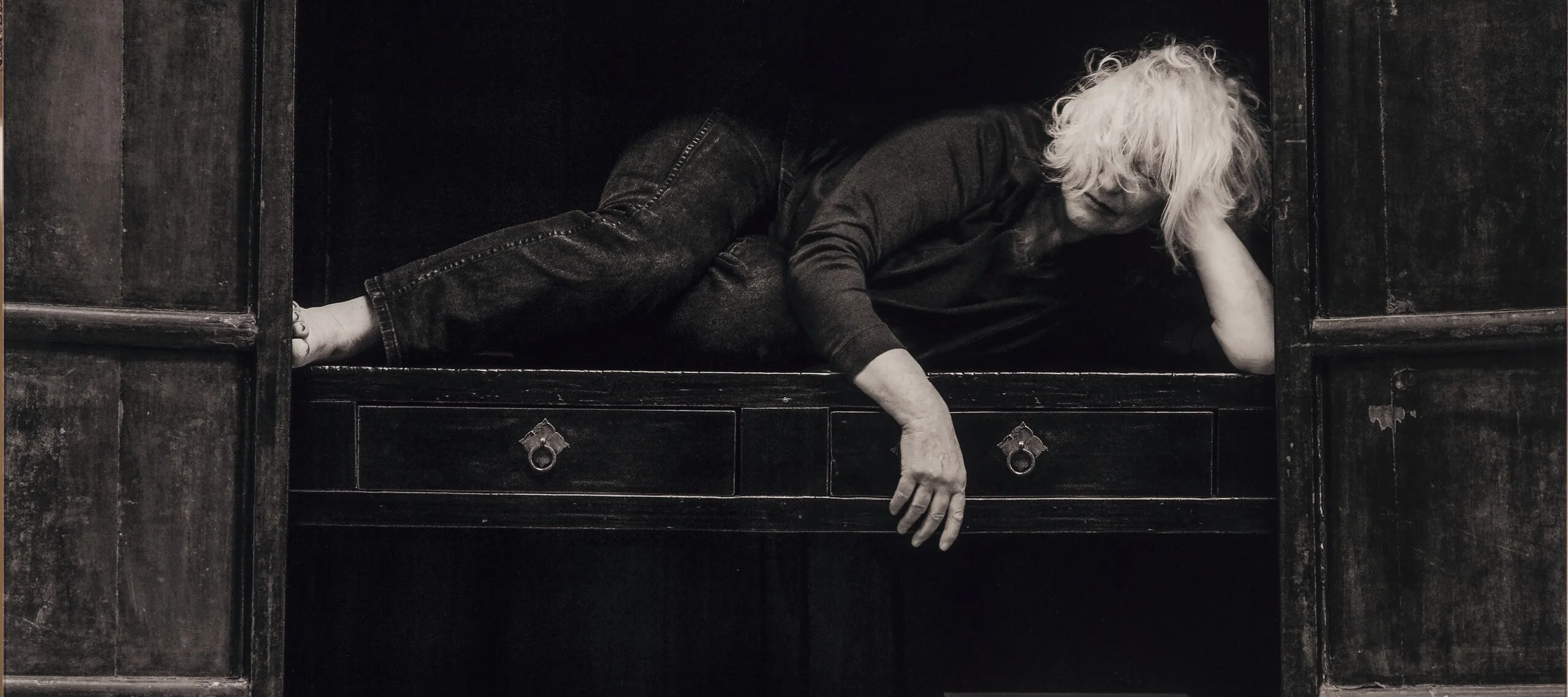 A black-and-white self-portrait of a light-skinned older woman with shoulder-length blonde hair who lays on the middle shelf of a large wooden cabinent. She is in a relaxed fetal position; one hand props her head up and the other is draped over the shelf. Above her a collection of tiny statues are arranged by size, on the shelf below her are a tea pot and two boxes.