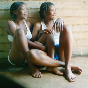 Two young black women sit closely together on the ground with arms and legs entwined suggesting an intimate relationship. They are looking off to the site with wide smiles.