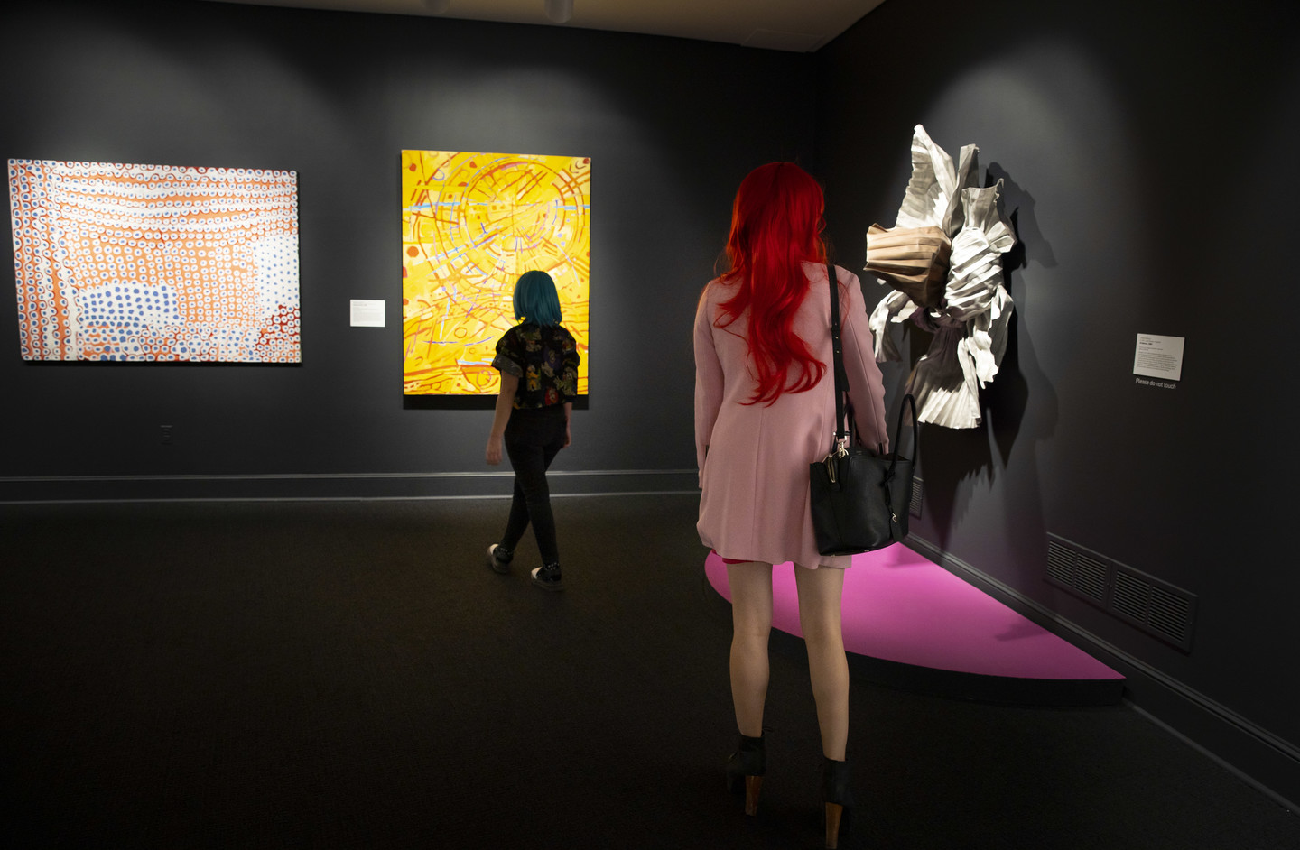 Corner gallery view of two visitors, one with long red hair and other with short blue-green hair looking at three artworks on dark walls.