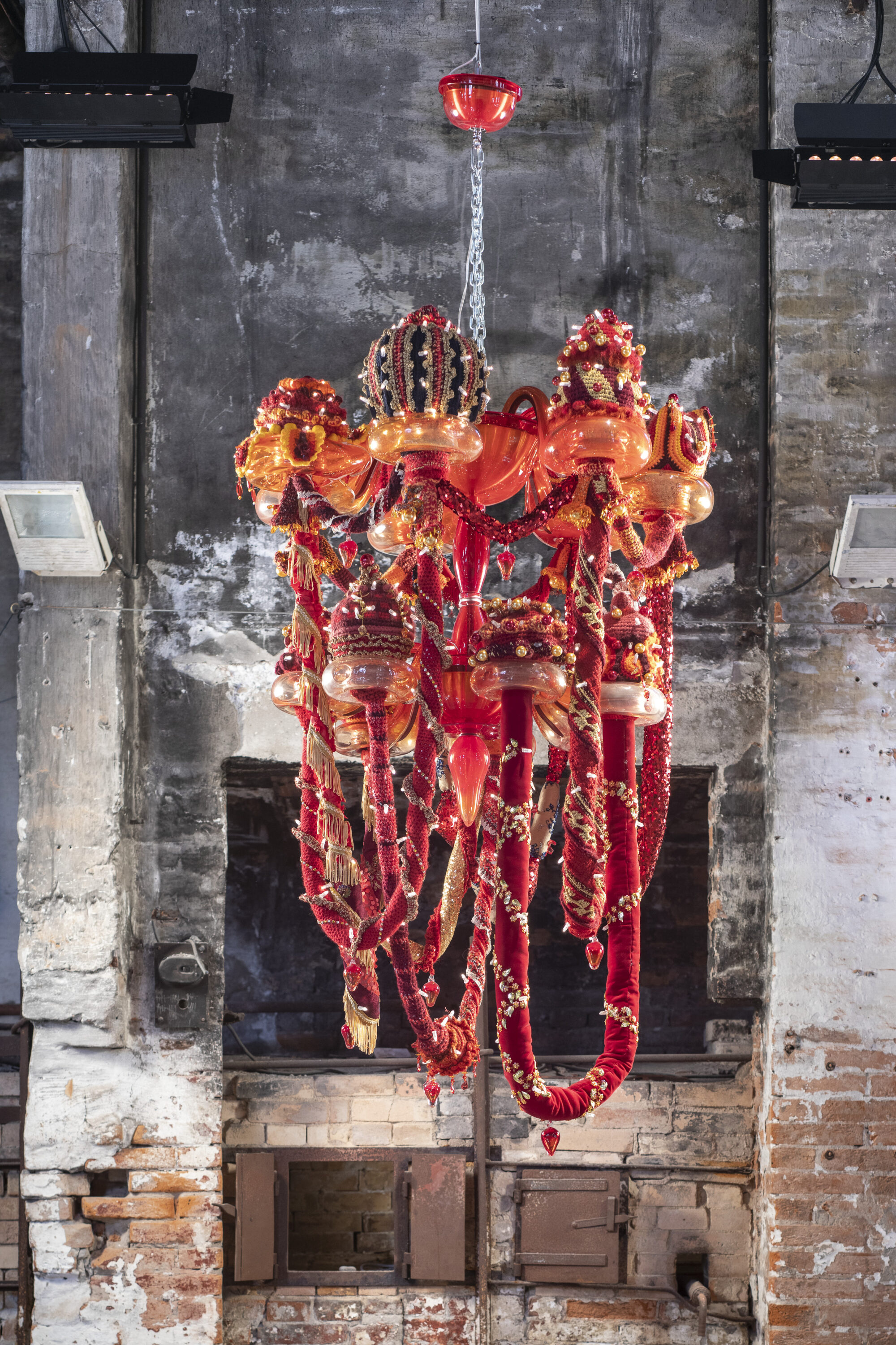 A large chandelier hangs from a chain in a brick room. The chandelier is made of red, orange, and yellow Murano glass, wool yarn, polyester fabrics, and LED lighting, with large red and gold elongated fabric garlands dangling at various heights, topped by embroidered crowns.