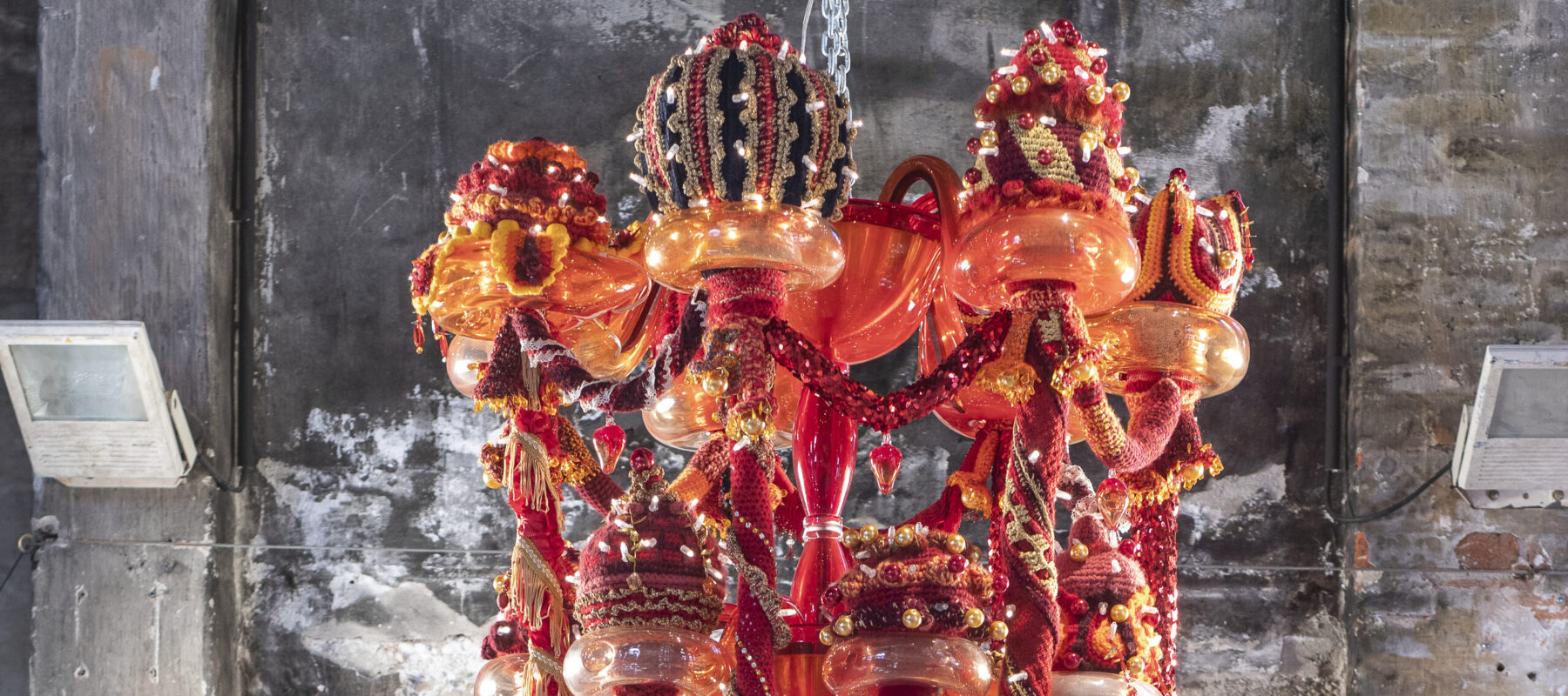 A large chandelier hangs from a chain in a brick room. The chandelier is made of red, orange, and yellow Murano glass, wool yarn, polyester fabrics, and LED lighting, with large red and gold elongated fabric garlands dangling at various heights, topped by embroidered crowns.