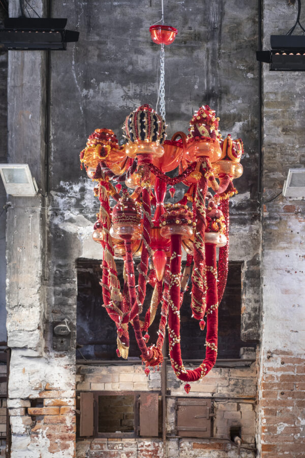 A large chandelier hangs from a chain in a room with white, gray, and red bricks. The chandelier is made of red, orange, and yellow Murano glass, wool yarn, polyester fabrics, and LED lighting.