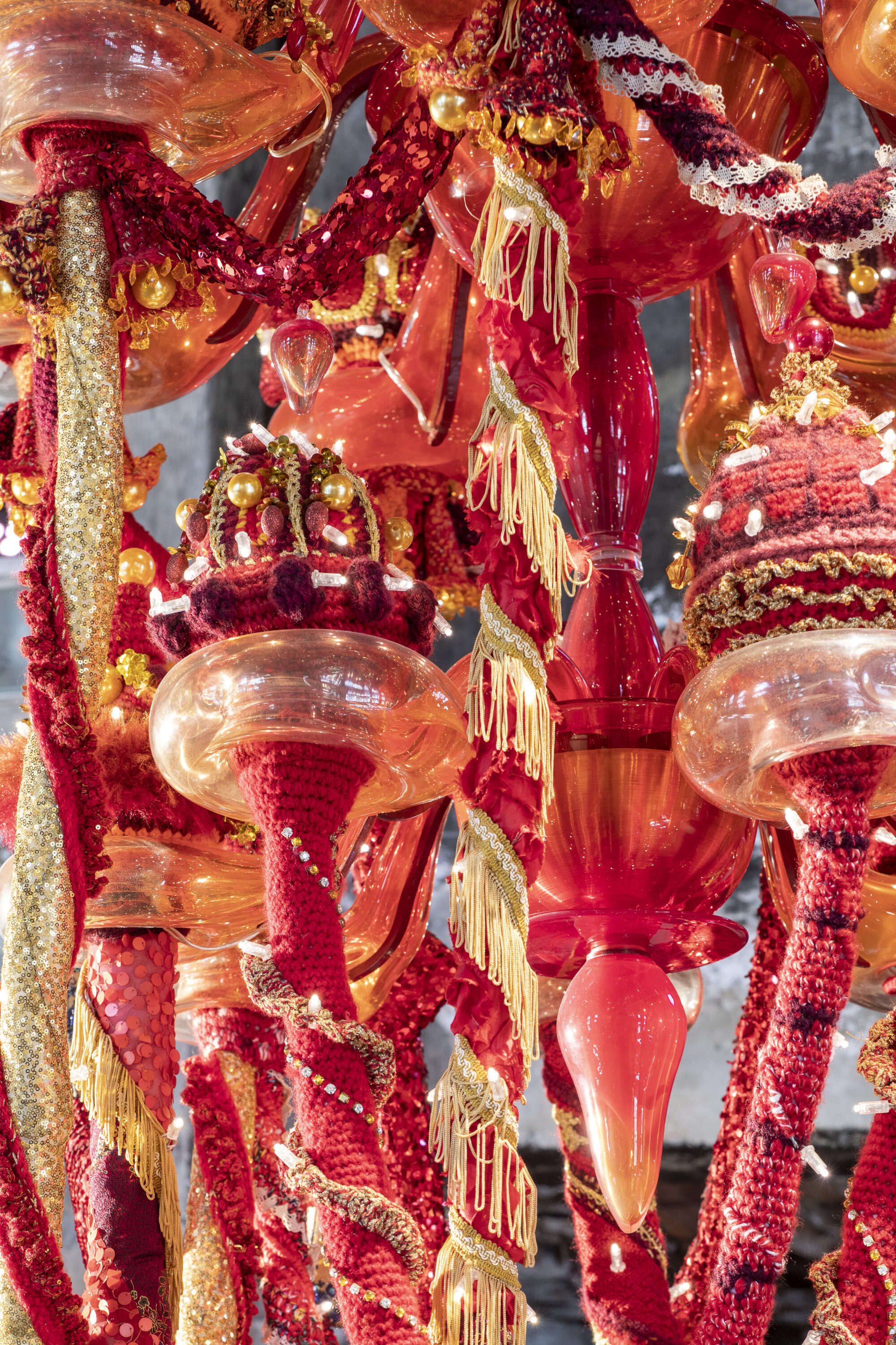 A color image of a detail of a chandelier made of red, orange, and yellow Murano glass, wool yarn, polyester fabrics, and LED lighting.