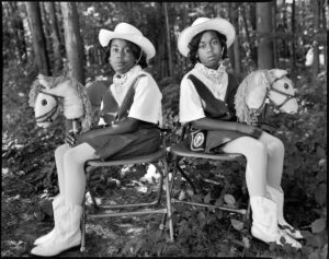 A black-and-white photograph of two dark-skinned young twins sitting in folding chairs against a woodsy background, facing the camera with their bodies angled outward in opposite directions. They wear matching cowgirl outfits, complete with hats, boots, and hobby horses.