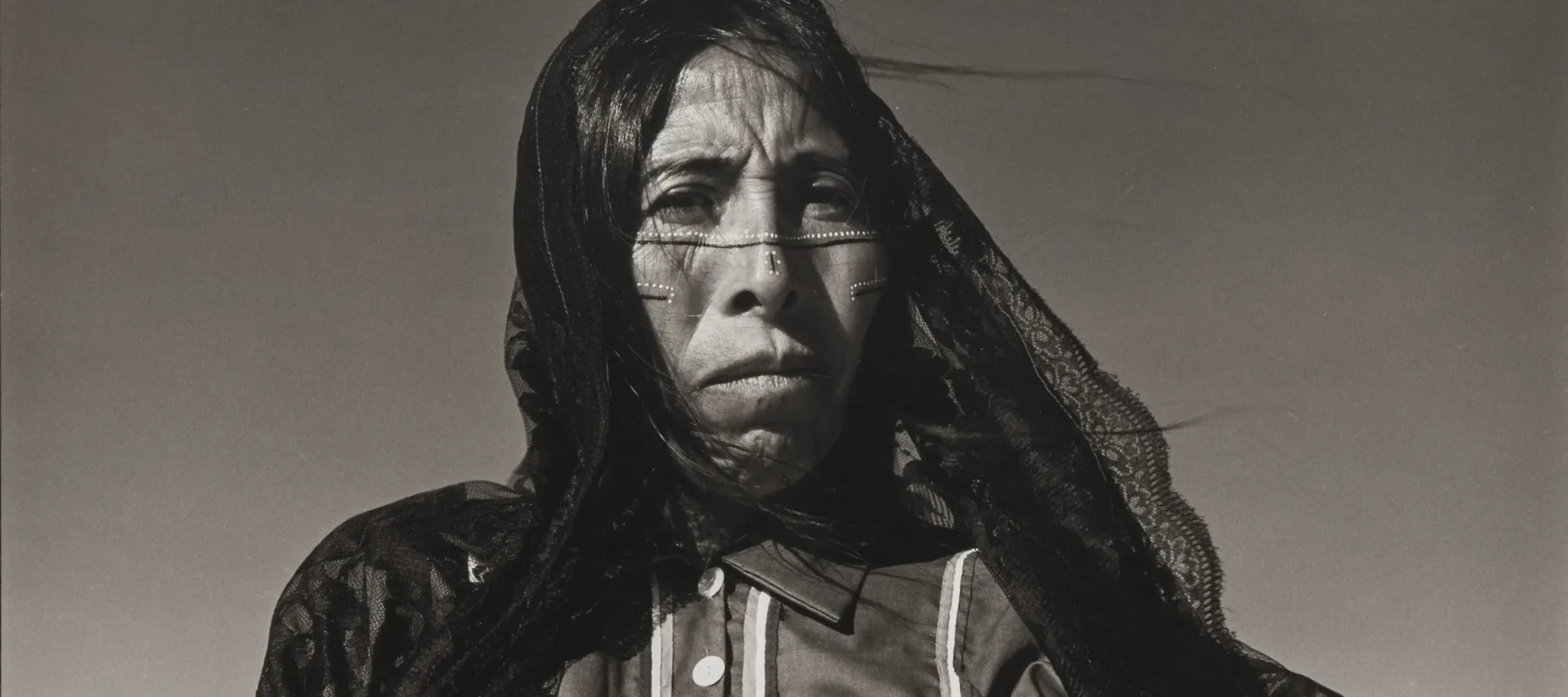 A black-and-white photograph of a Seri woman standing against a clear sky with medium-toned skin and dots and lines marking her face. Wisps of her long, dark hair and a long, lacy, dark veil are caught in the breeze. She wears a collared, button-up shirt.