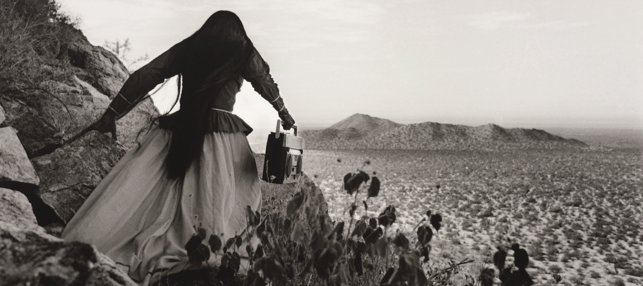 A black-and-white photograph shows the back of a woman as she crests a rocky path above a vast desert landscape beneath an expansive sky. Her traditional, ethnic full skirt, long-sleeved blouse, and long, straight, dark hair contrasts with the modern portable stereo she carries.