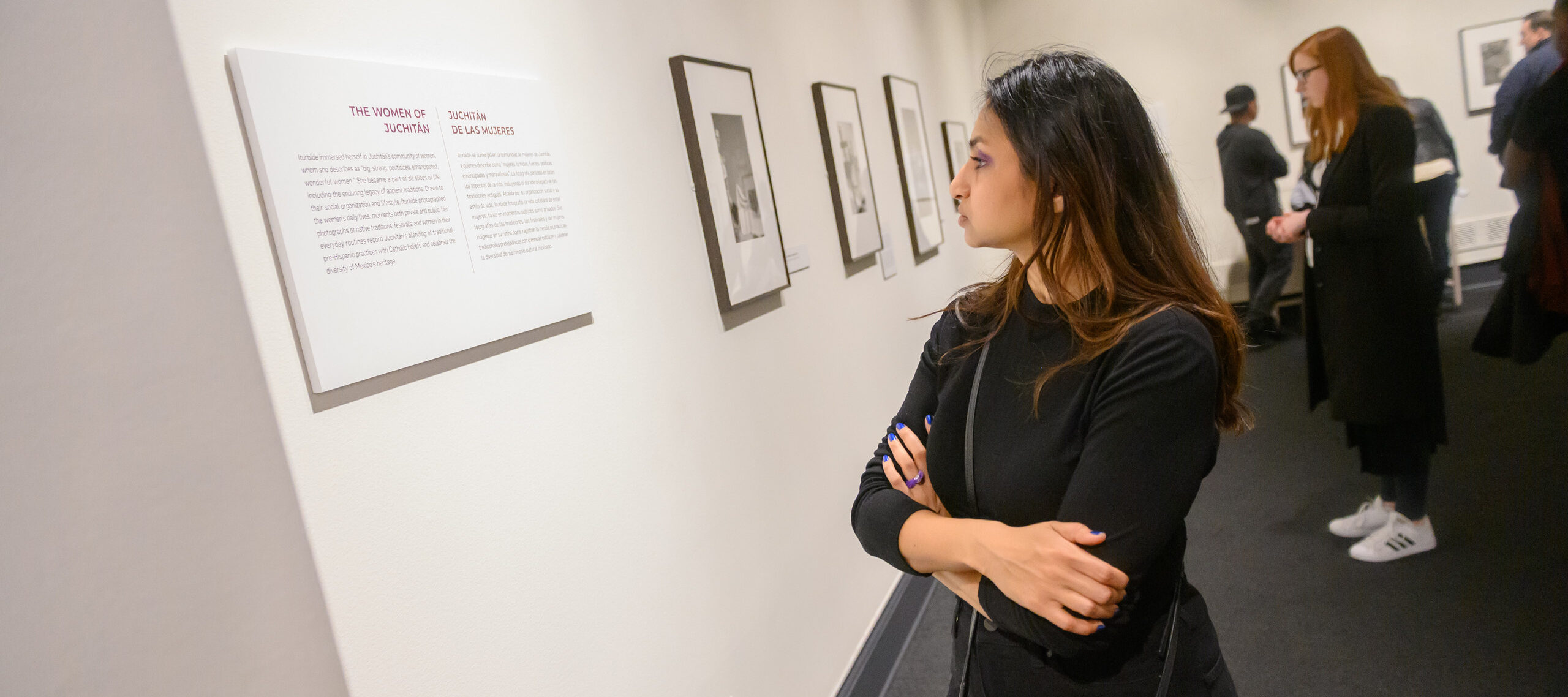 A medium light-skinned young woman in a black top crosses her arms while reading a gallery exhibition label intently.