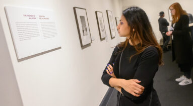 A medium light-skinned young woman in a black top crosses her arms while reading a gallery exhibition label intently.