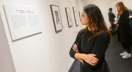 A medium light-skinned young woman in a black top crosses her arms while reading a gallery exhibition label intently.