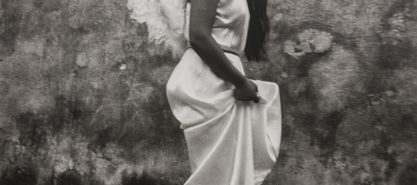 A black-and-white photograph of a woman in profile from the shoulders down with feathery white wings on her back walking and hoisting up her silky white dress.