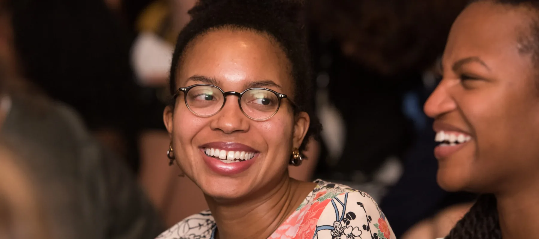 Two dark-skinned adults grin together amid an audience of people. One adult faces the viewer and wears glasses, a patterned top, and their dark hair in a bun; while the other is in profile and and has long, dark braids.