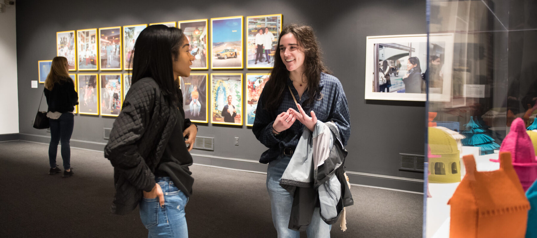 Two young women, one with medium-dark skin and dark hair and one with light skin and dark hair stand in front of a display case, chatting.