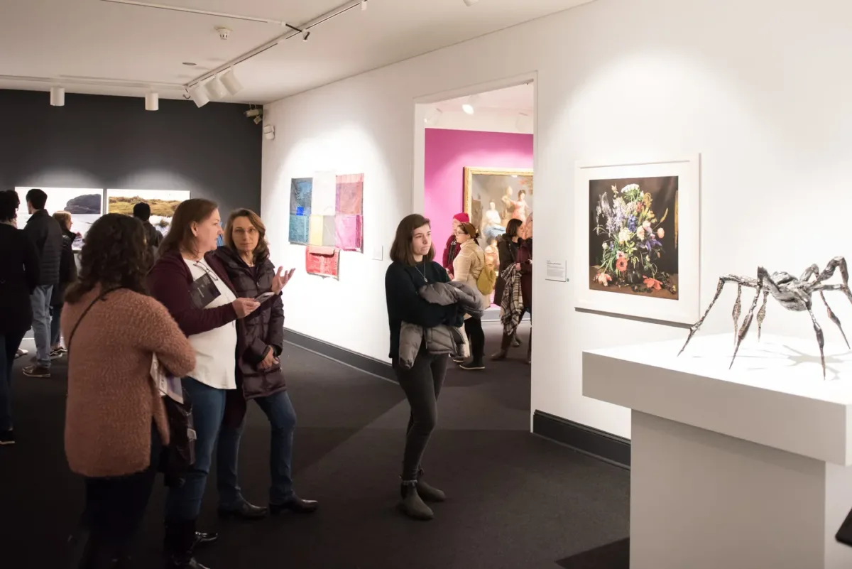 Gallery view of group of women looking at a sculpture of a black spider that stands on a high white platform.