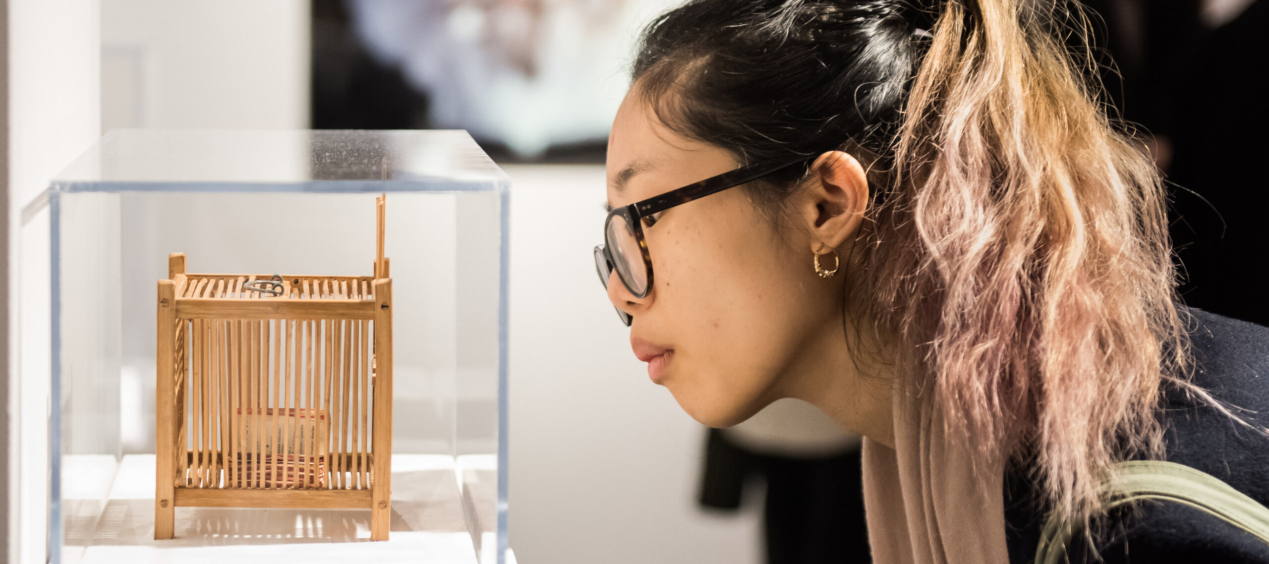 Partial gallery view of a young woman in a ponytail and glasses bending down to look at a tiny contemporary sculpture in a glass vitrine.