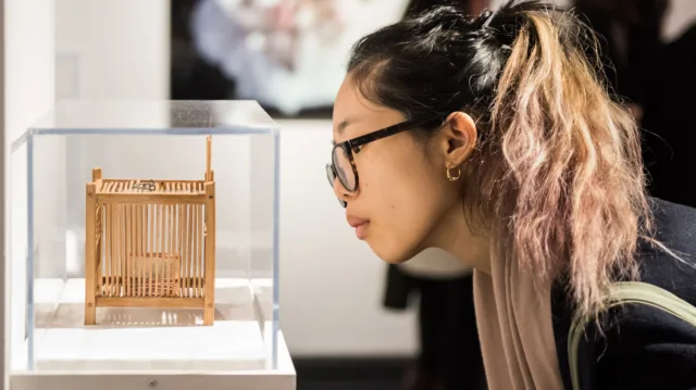 Partial gallery view of a young woman in a ponytail and glasses bending down to look at a tiny contemporary sculpture in a glass vitrine.