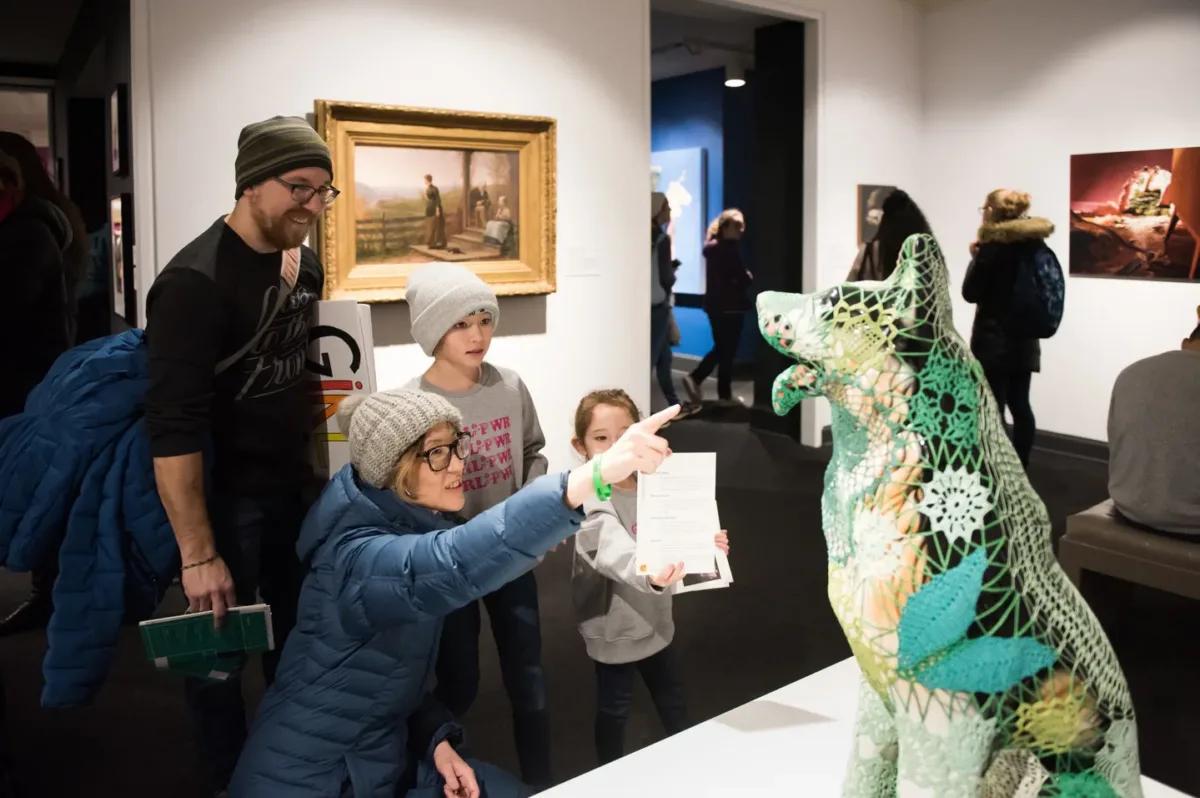 Gallery view of visitors looking at a contemporary sculpture of a dog covered in green knitting in the foreground. A woman kneels pointing at the dog, while a man and two children stand next to her