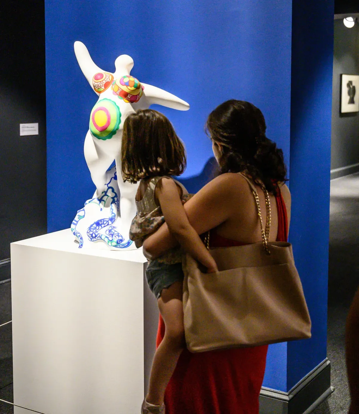 A medium-skinned woman holds a young child while both look at a voluptuous, faceless, white statue that stands on a white pedestal.