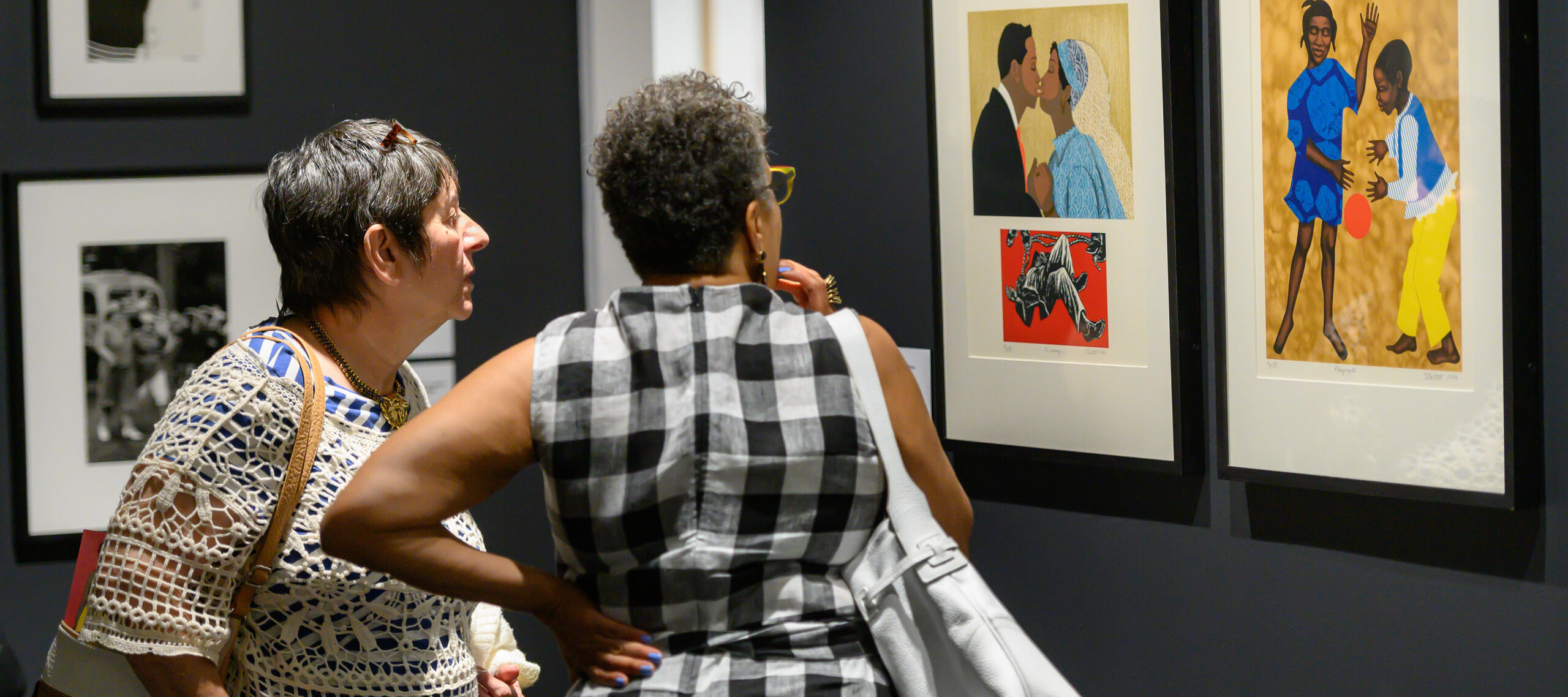 Two older women, one light-skinned with gray hair and one medium-dark skinned with short dark hair, stand next to one another deep in conversation about a series of prints by Elizabeth Catlett.