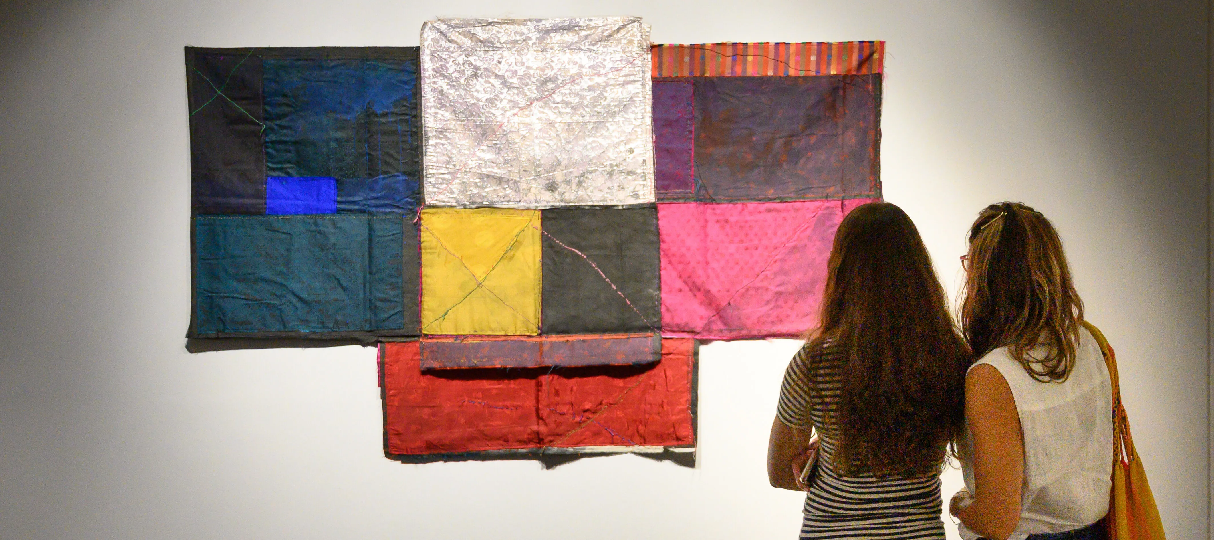 Two people standing next to each other in casual clothes with long hair seen from behind; they are looking at a t-shaped, colorful textile collage artwork hung on a white gallery wall.