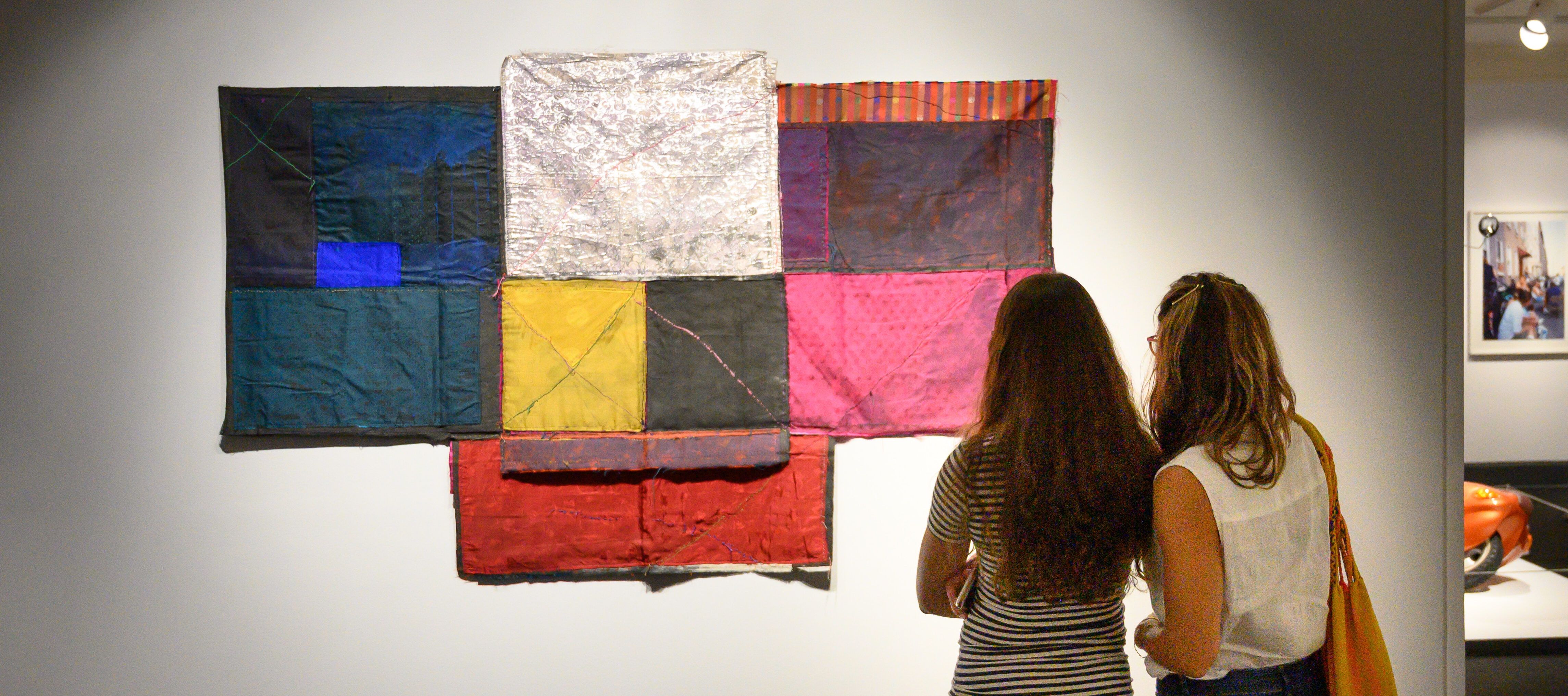 Two people standing next to each other in casual clothes with long hair seen from behind; they are looking at a t-shaped, colorful textile collage artwork hung on a white gallery wall.