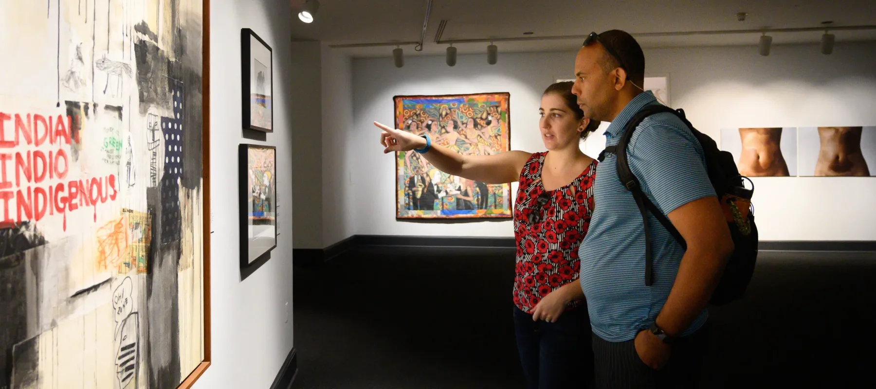 Partial gallery view of a man and woman pointing at framed artworks on a wall. The large work in the foreground has the words “INDIAN INDIO INDIGENOUS” in red hand-drawn letters against a background of red and black painted sections and drips.