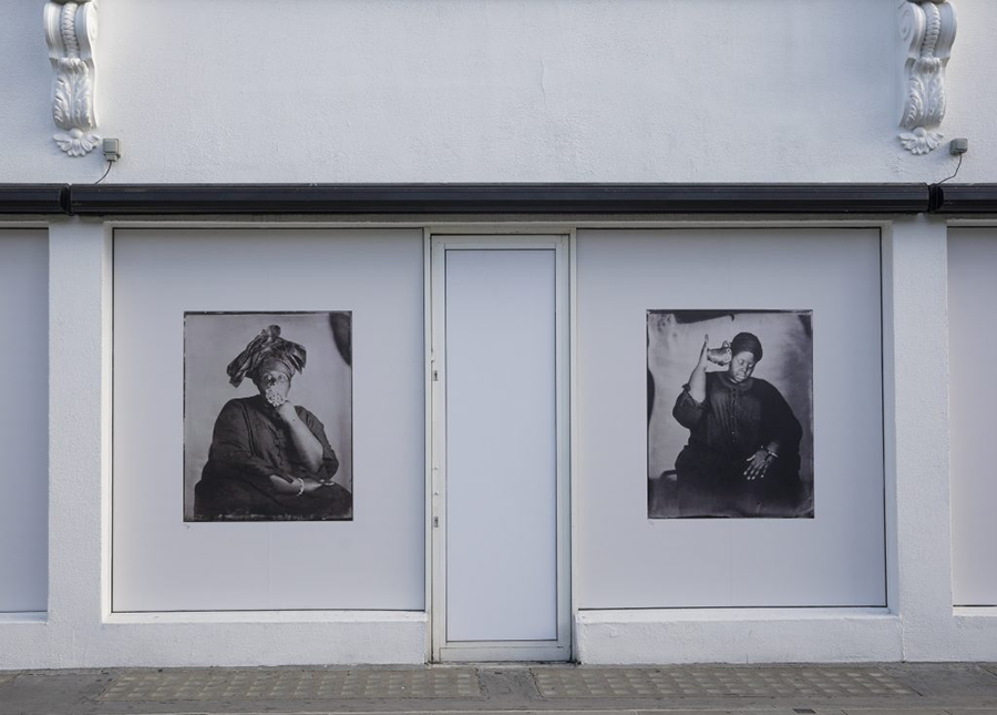 Two poster-size black and white photographs, each an artful portrait of a dark-skinned woman with her hand to her face in one, and holding a shell to her ear in the other, occupy the façade of a low white city building. 