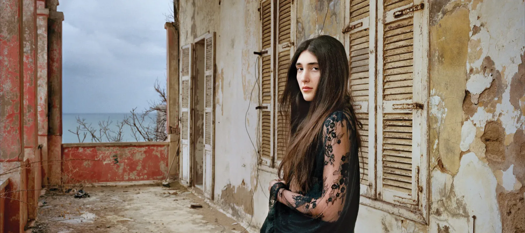 A light-skinned young woman with long, dark brown hair in a black, long lace sleeved dress stands confidently in a crumbling loggia. She gazes at the viewer with a serious, captivating look.