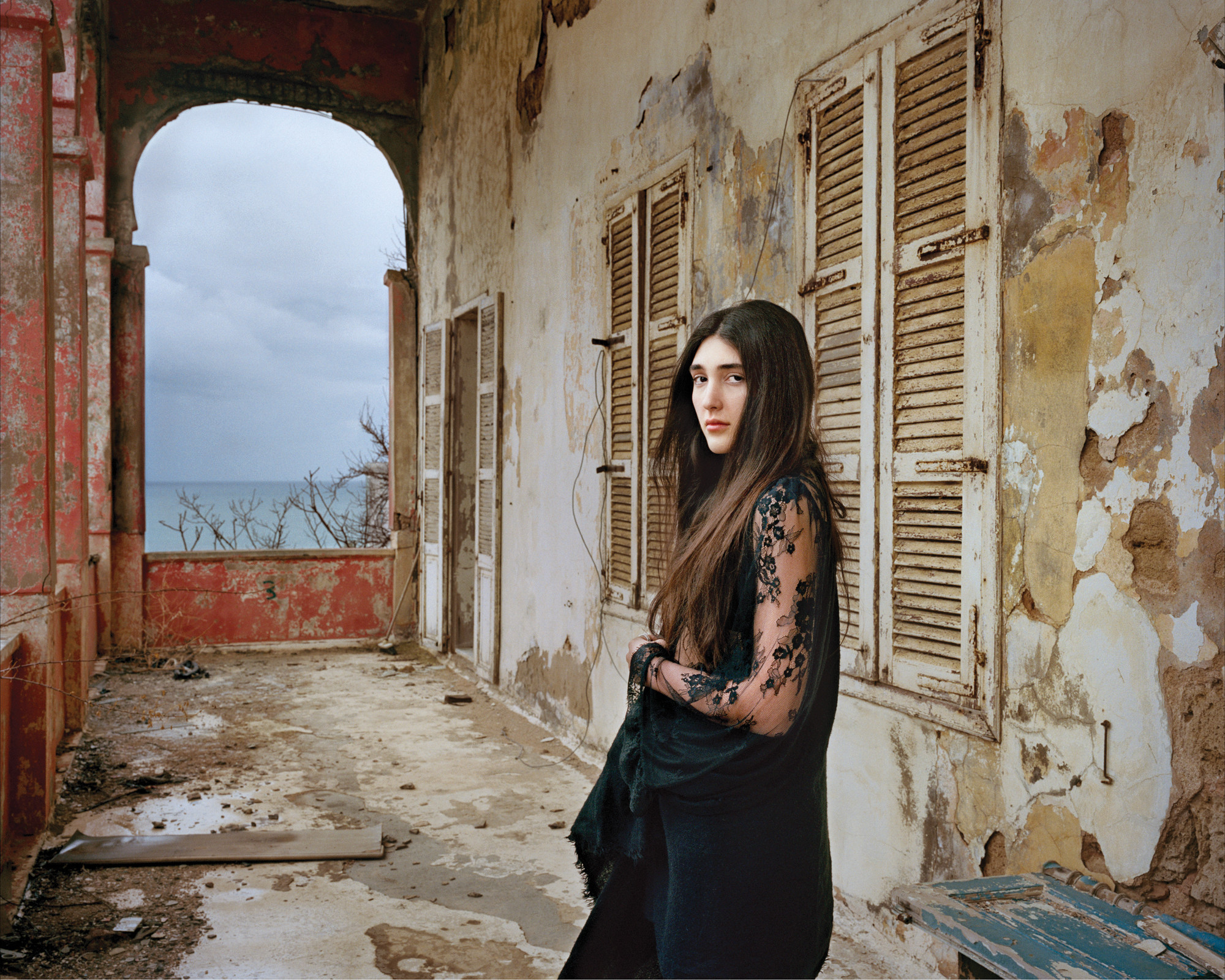 A light-skinned young woman with long, dark brown hair in a black, long lace sleeved dress stands confidently in a crumbling loggia. She gazes at the viewer with a serious, captivating look.