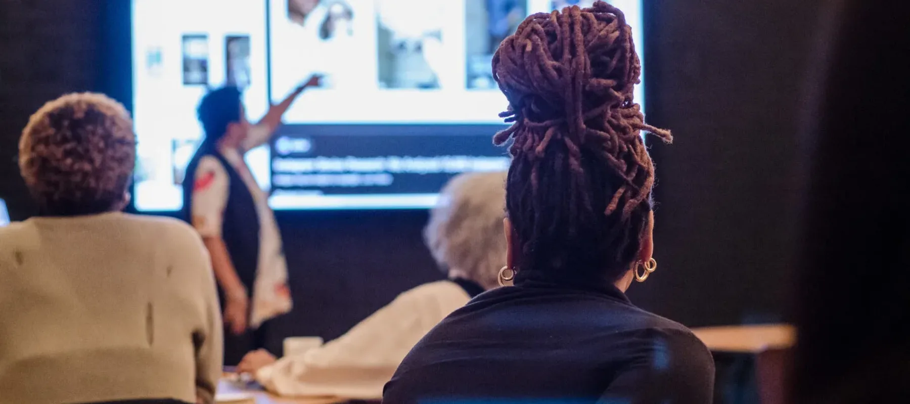 A variety of people sitting at tables and seen from behind. They all face forward toward a presenter speaking and pointing at a presentation being projected on the wall.