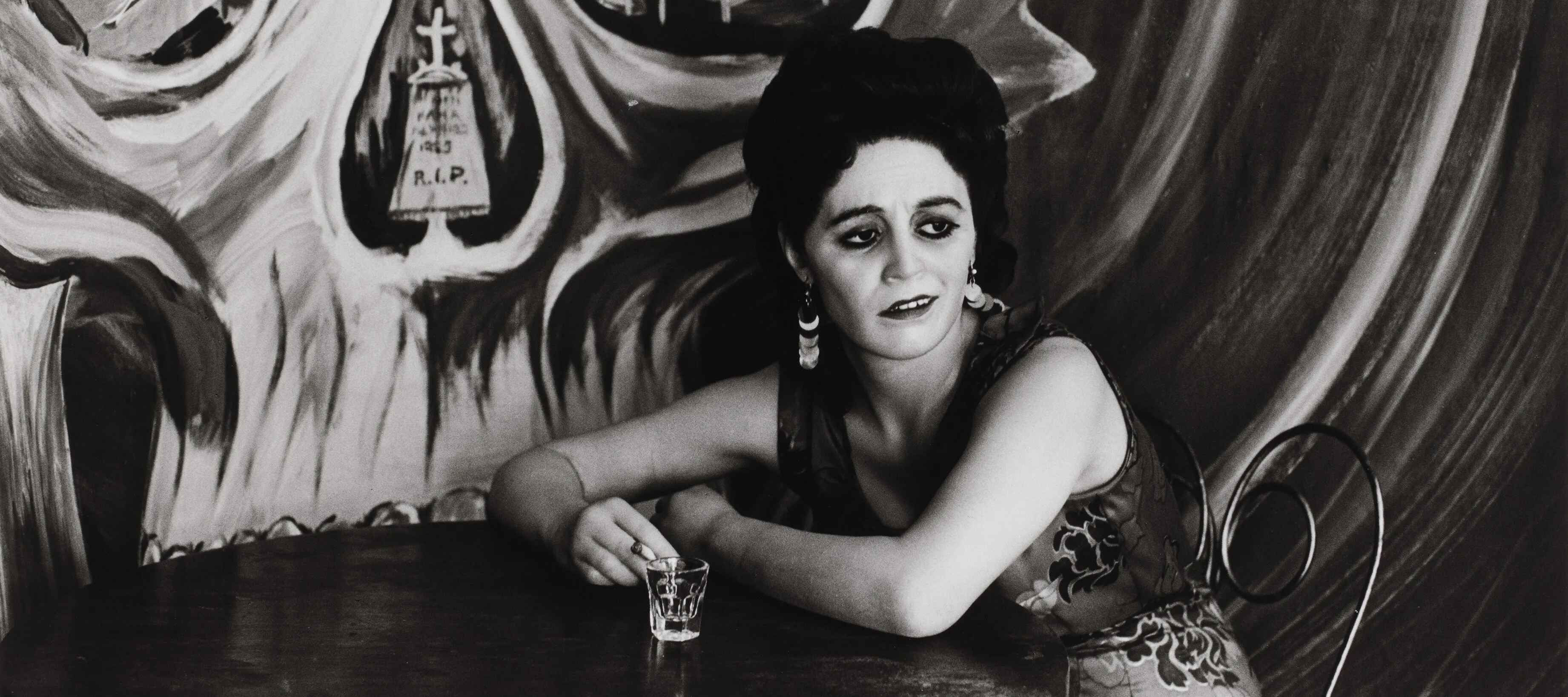 Black and white photograph of light skin toned woman with long dark hair sitting at a cafe table. On the wall behind her is a painted mural of a large skull with hospital beds visible in the eye sockets and a tombstone in the nasal cavity.