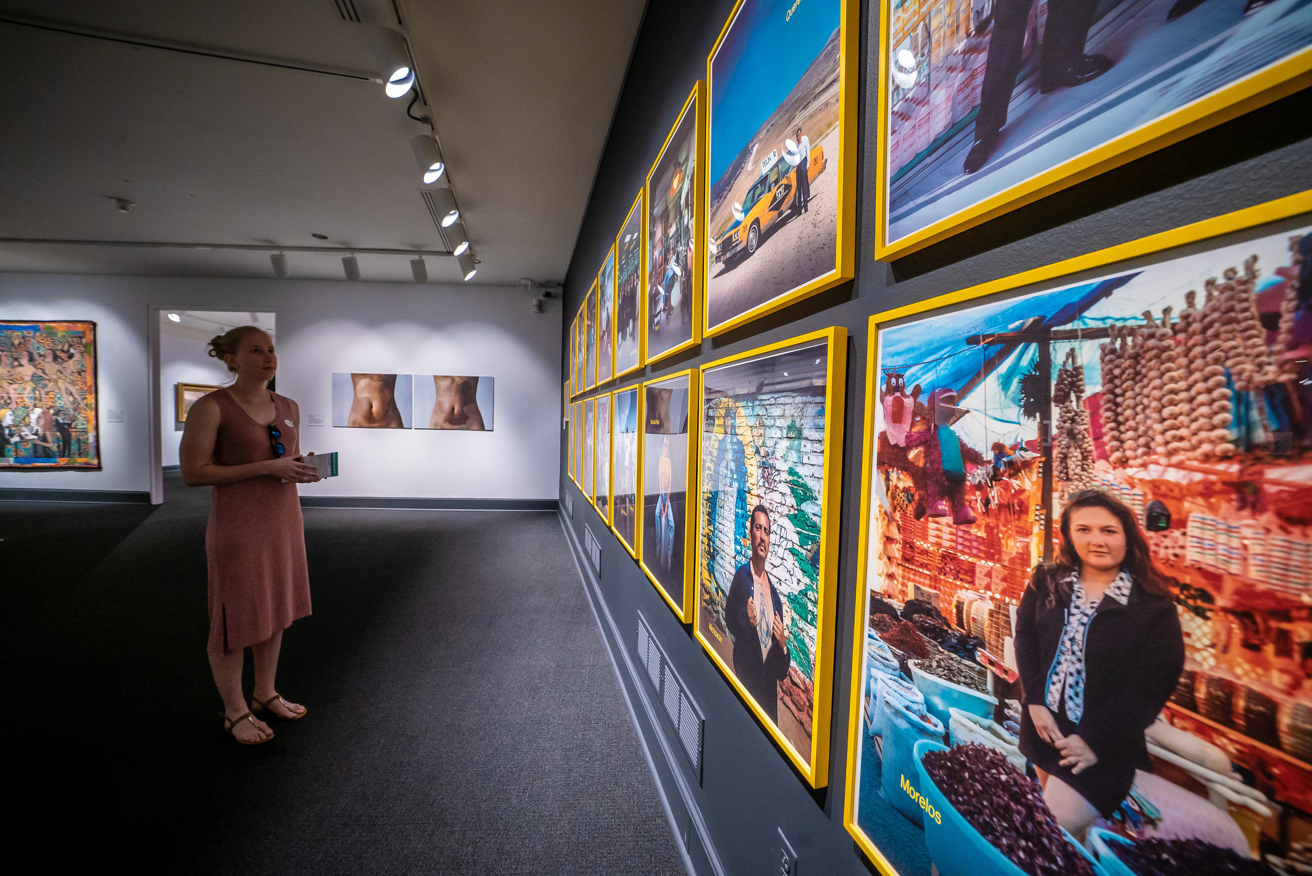 Gallery view of a person with light skin looking at a wall of photographs in bright yellow frames. Two artworks hand on the far wall.