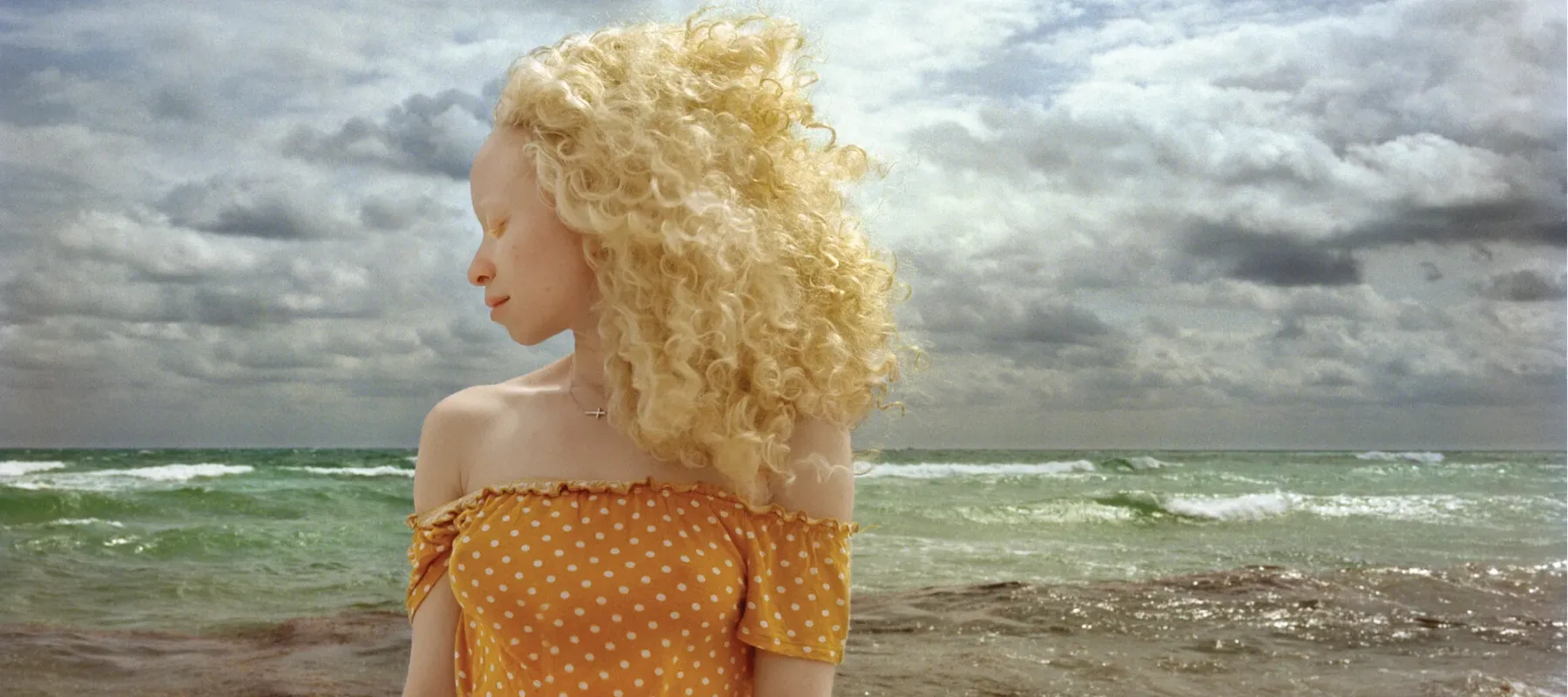 In front of a stormy ocean, a woman with light skin and blonde, wind-blown hair stands with head turned in profile and eyes closed.