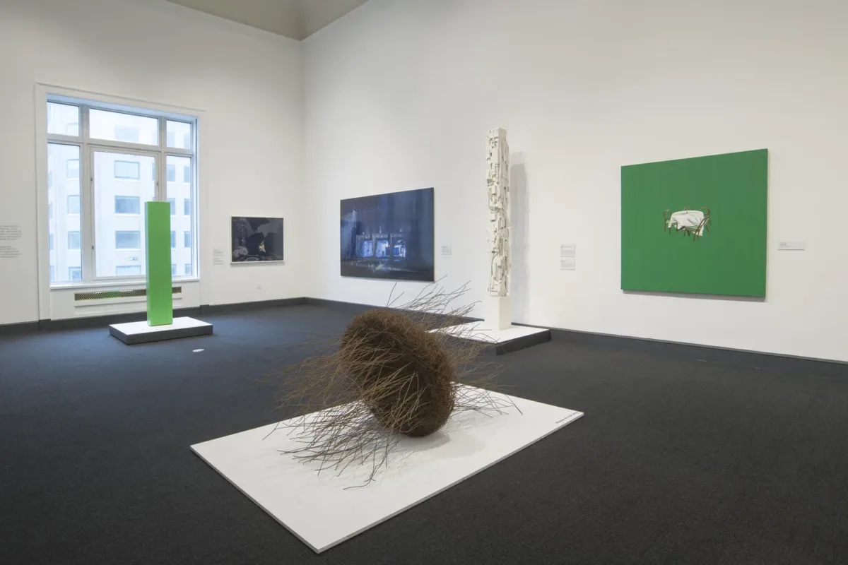 Partial view of a gallery with six contemporary artworks, including painting of small chairs and table with white tablecloth centered on a neon green background, two photographs on adjacent walls, three sculptures rest on white platforms on the floor.