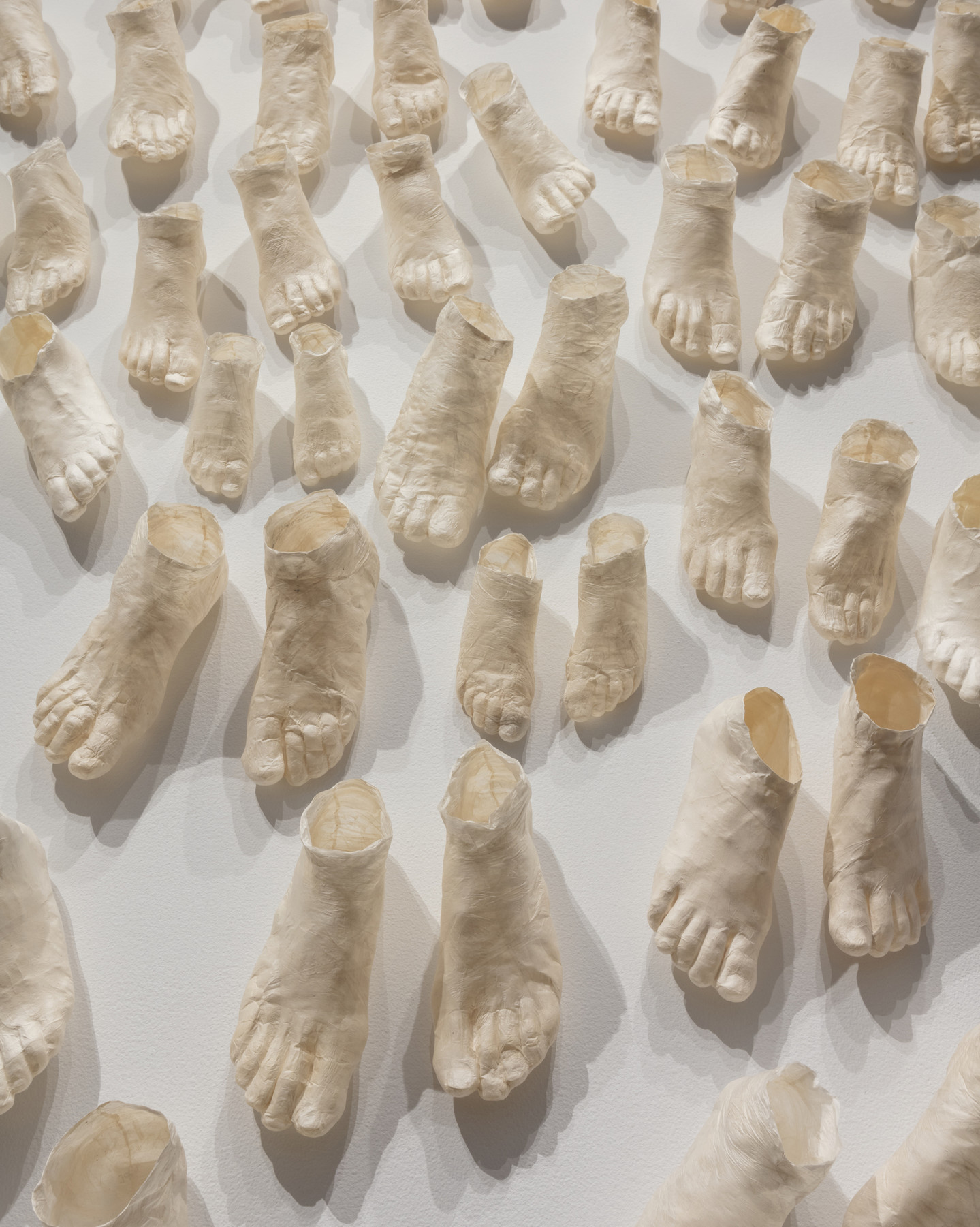 Dozens of pale, hollow, ghostly feet of different sizes made of translucent paper arrayed in pairs on a white background.