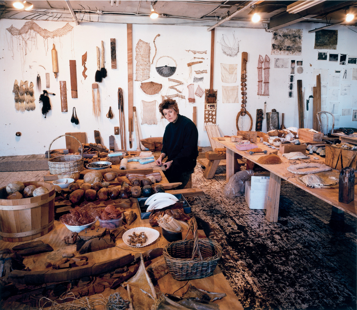 A light-skinned woman with close-cropped hair stands between two large plywood tables, looking up from her work. The tables and walls are filled with natural objects and artworks, including hanging weavings, spools of thread, carved wooden rocks, and wicker baskets.