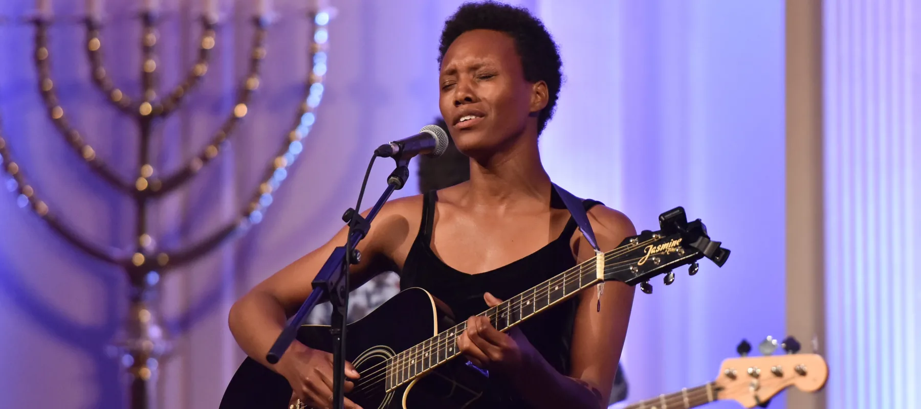 A dark-skinned adult woman in a black tank-top and pants with short, curly hair plays a black guitar and sings into a microphone while standing against a light purple background.