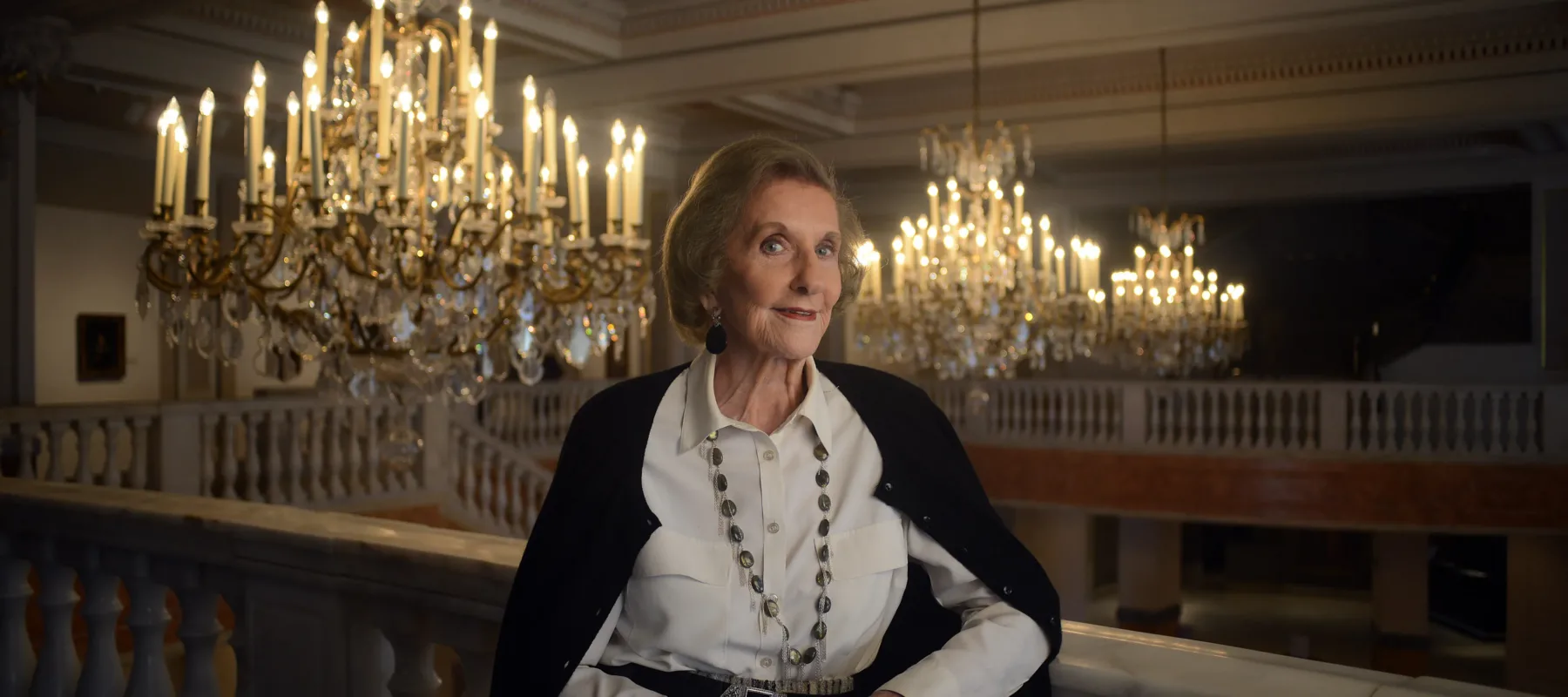 A light-skinned older woman in a collared white shirt and black cardigan leans against a railing with a slight smile. Ornate chandeliers can be seen behind her.