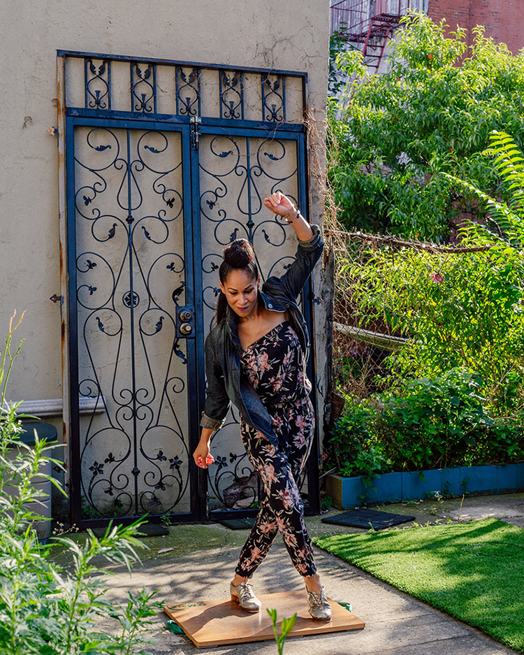 A dark-skinned woman wearing a black and pink floral jumpsuit, a dark denim jacket, and metallic shoes dances on a wooden plank next to a small patch of grass. Her legs are crossed, and one arm is swinging up over her head; she is clearly moving quickly, and enjoying herself.