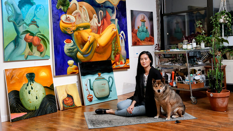In a room with hardwood floors, large windows and white walls, a light-skinned woman sits on a small carpet with a brown dog. Behind them, large, colorful and surreal paintings take up the entire wall, and houseplants mingle with a steel cart of art supplies in the corner.
